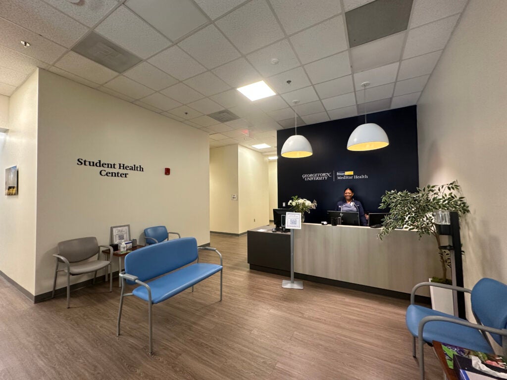 New waiting room in the entrance of the Student Health Center. Features chairs, a navy blue accent wall with branding, and overhead lighting.